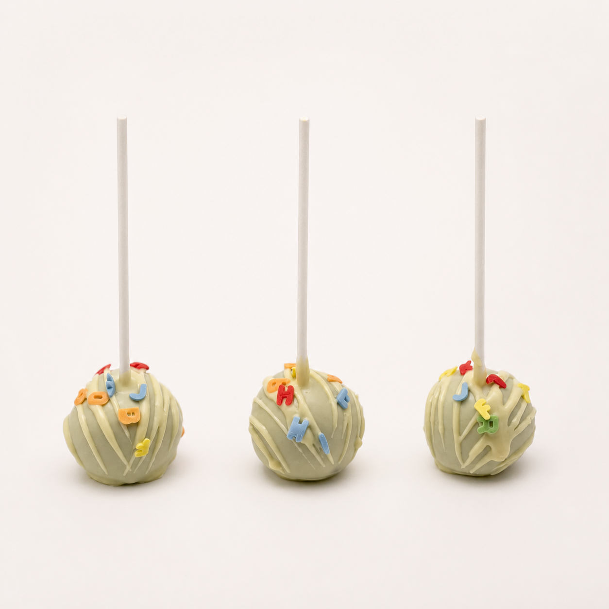 Einschulungs-Cake-Pops – Produktfoto Drei grüne Einschulungs-Cake-Pops mit weißer Schokolade und bunten Buchstaben vor fast weißem Hintergrund